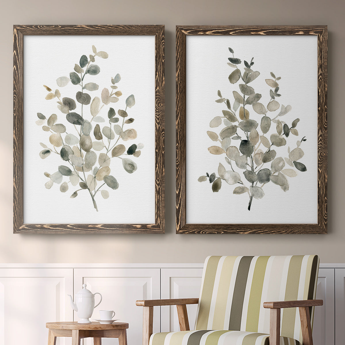 Set of two walnut barnwood framed canvas prints depicting two minimalist watercolor botanical studies featuring delicate eucalyptus branches with rounded leaves above a sideboard in a cozy sitting room.