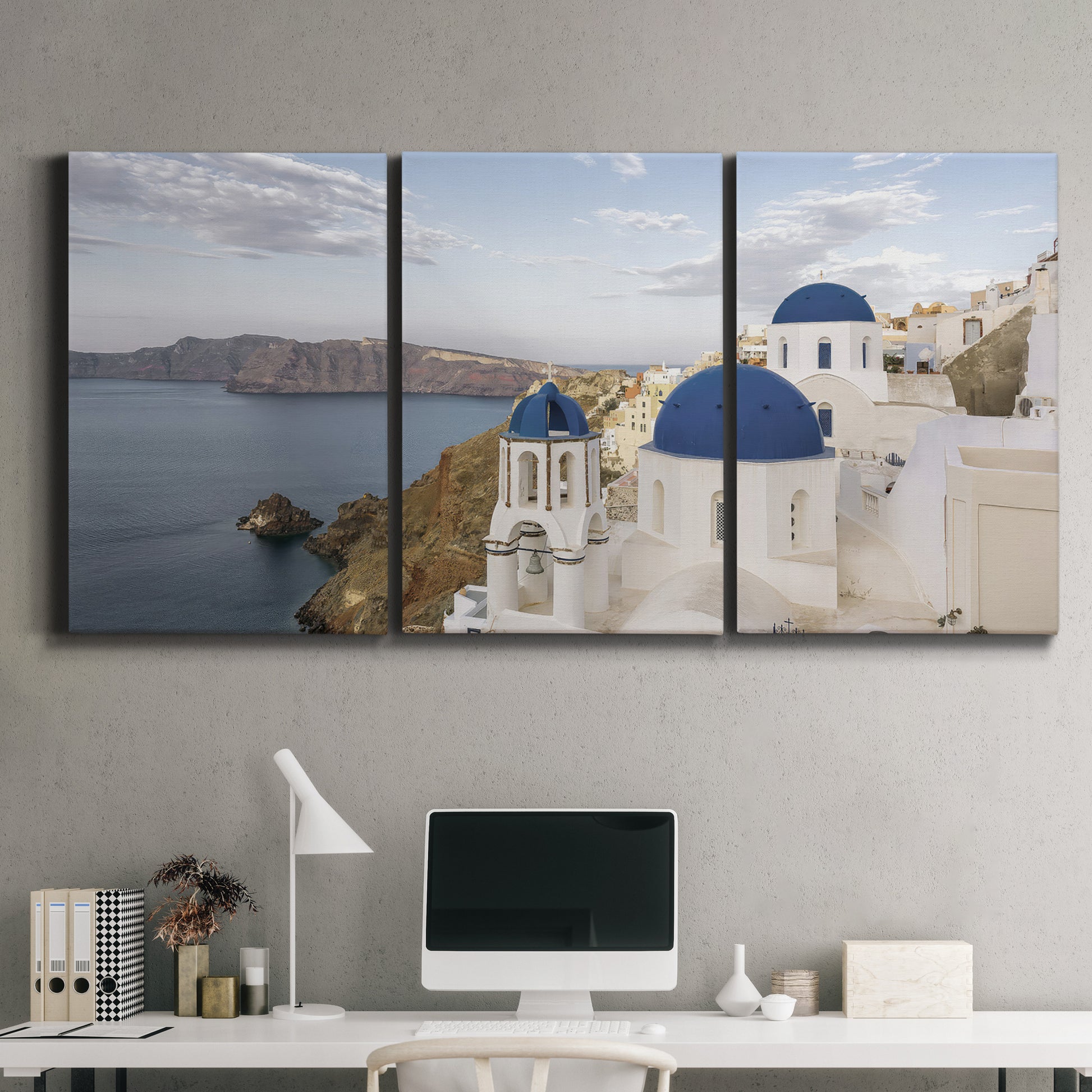 Three-panel canvas wall art featuring white buildings overlooking sea cliffs, displayed above a desk in a home office.