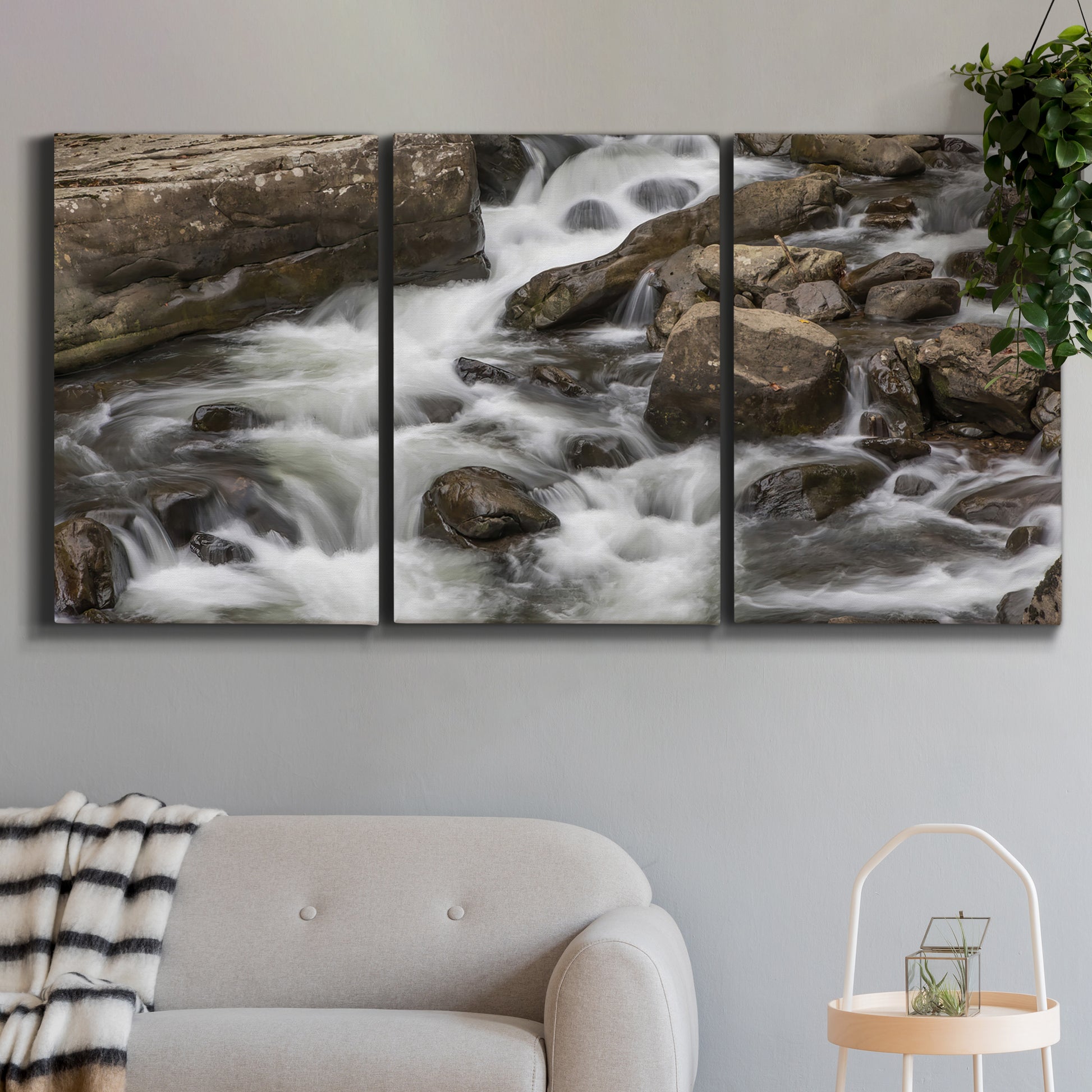 Three-panel canvas wall art featuring rushing river water over rocks, displayed in a modern living room above a sofa.