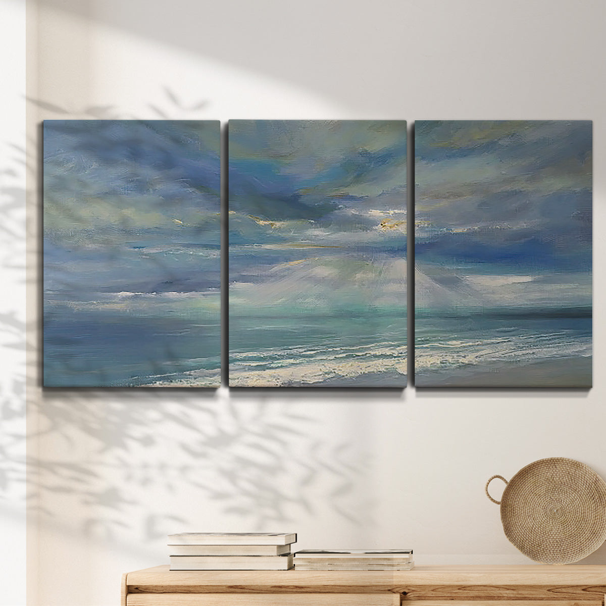 Three-panel canvas wall art featuring sunbeams breaking through coastal clouds, displayed in a minimalist living room above a console table.