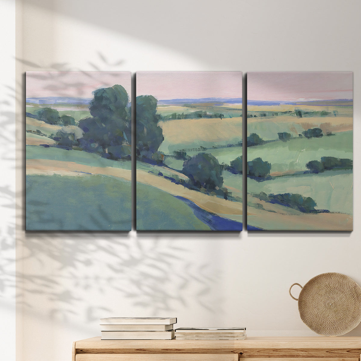 Three-panel canvas wall art featuring pastoral countryside with rocky outcrop, displayed in a minimalist living room above a console table.