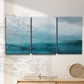 Three-panel canvas wall art featuring vertical coastal elements with rising light, displayed in a minimalist living room above a console table.