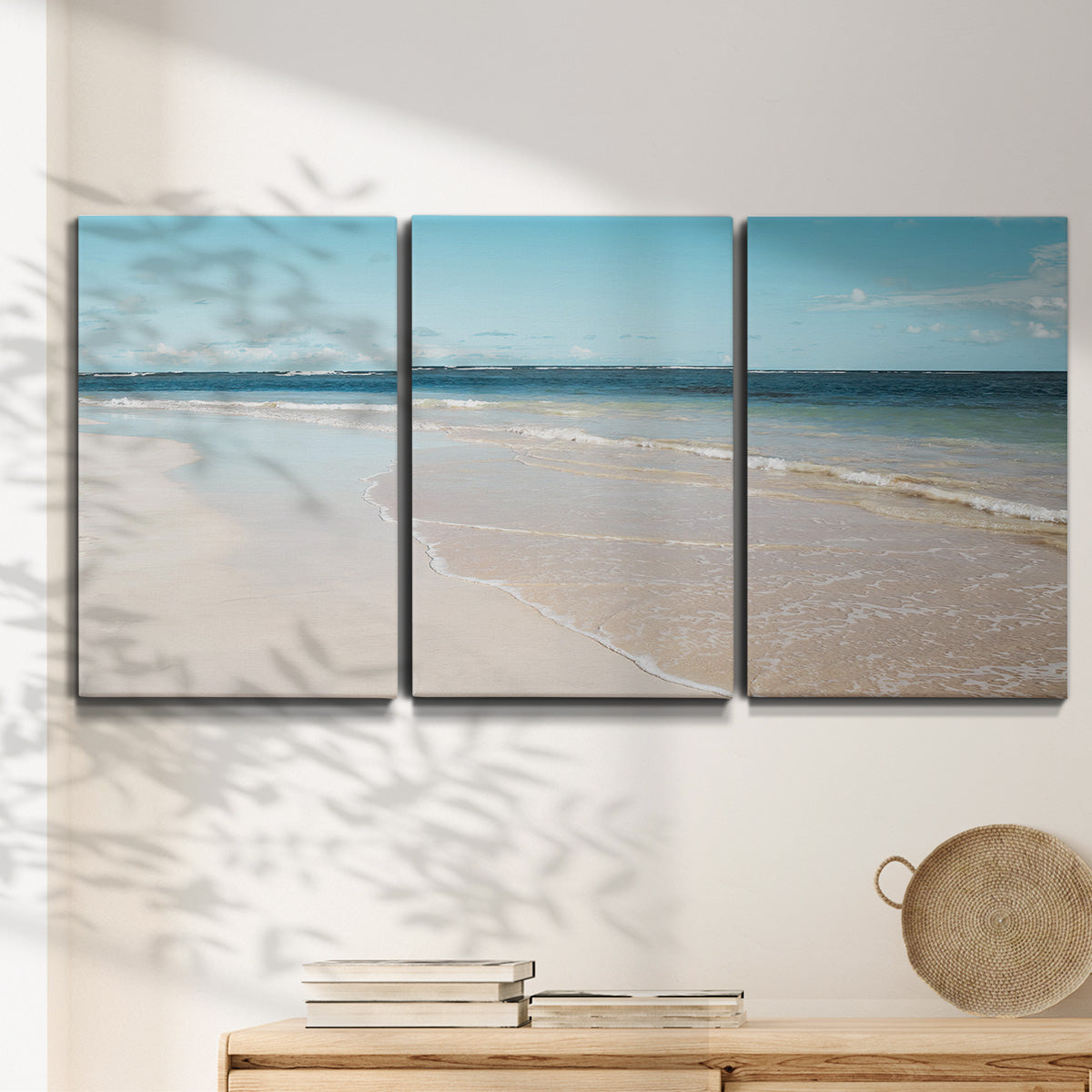 Three-panel canvas wall art featuring white sand beach with gentle waves, displayed in a minimalist living room above a console table.