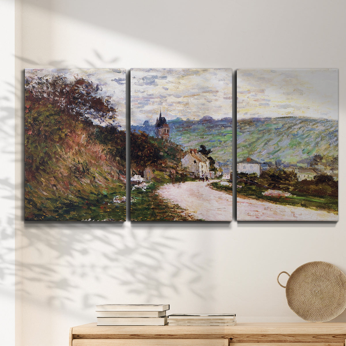 Three-panel canvas wall art featuring country road through village, displayed in a minimalist living room above a console table.