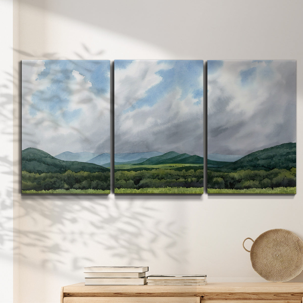 Three-panel canvas wall art featuring pastoral mountain landscape in blue, displayed in a minimalist living room above a console table.