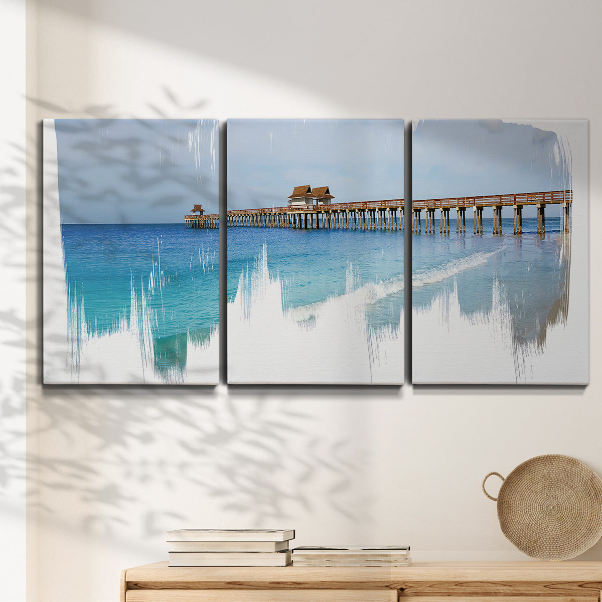 Three-panel canvas wall art featuring coastal pier extending over water, displayed in a minimalist living room above a console table.