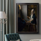 Quiet interior genre scene of a woman with scales by Johannes Vermeer in an ornate black frame with gilded inner detail displayed in a neutral interior setting.