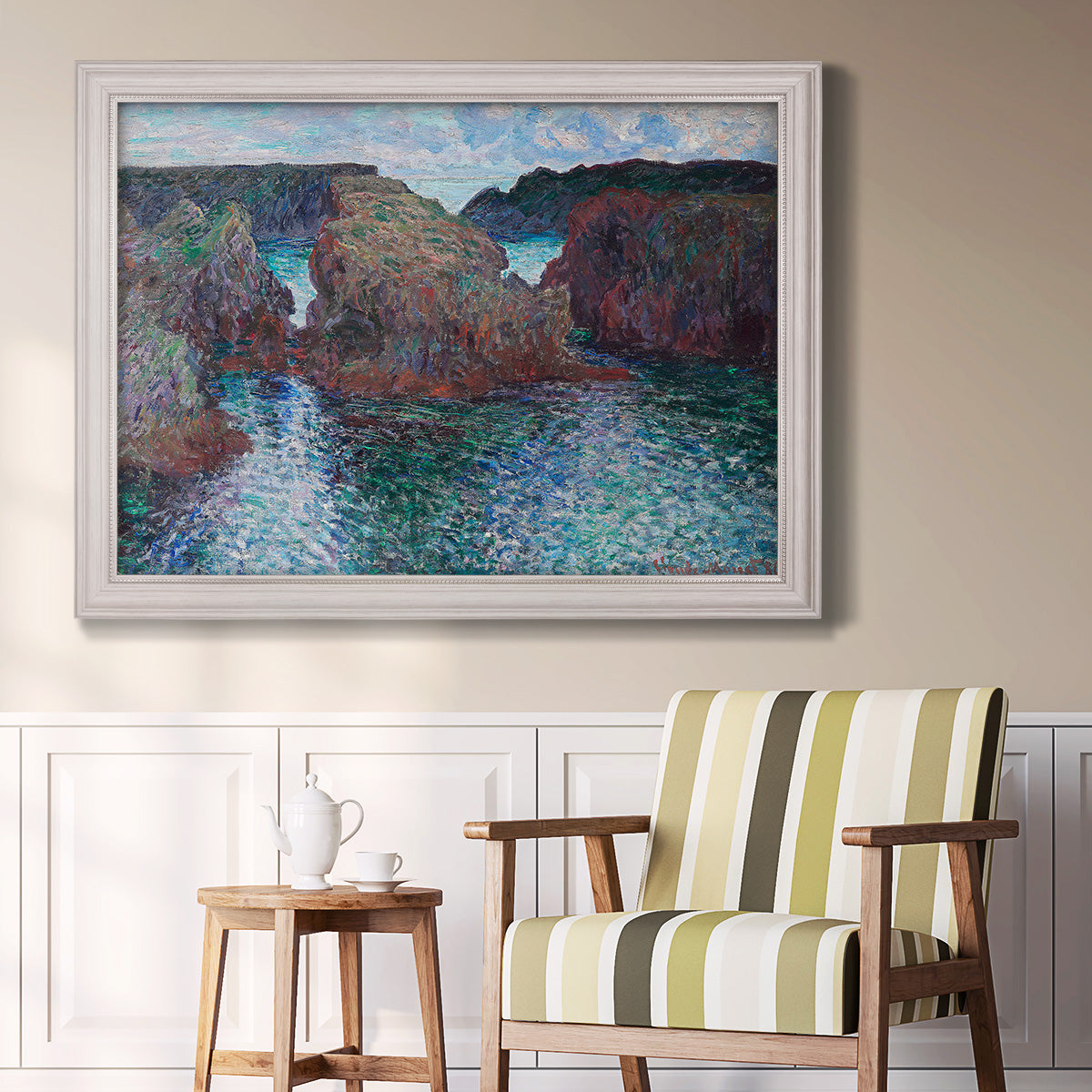 Impressionist rocky seascape by Claude Monet in an ornate weathered white frame displayed in a light-toned interior setting.