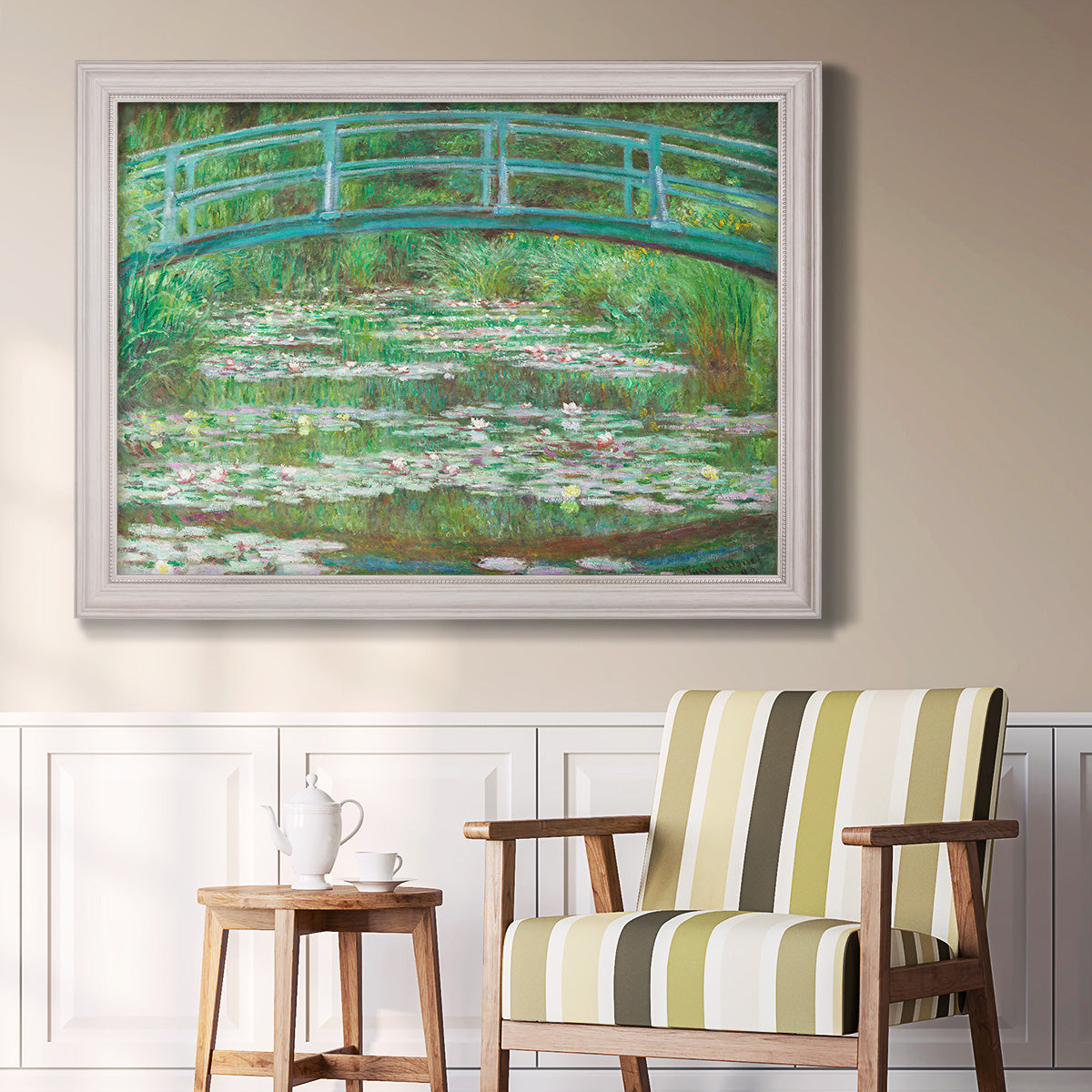 Impressionist garden scene with Japanese bridge and water lilies by Claude Monet in an ornate weathered white frame displayed in a light-toned interior setting.