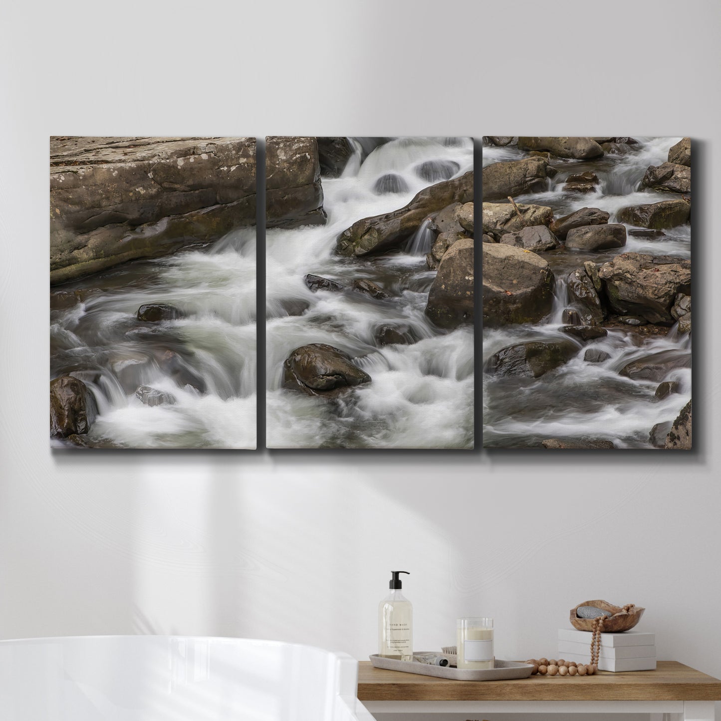 Three-panel canvas wall art featuring rushing river water over rocks, displayed in a bathroom above a bathtub.