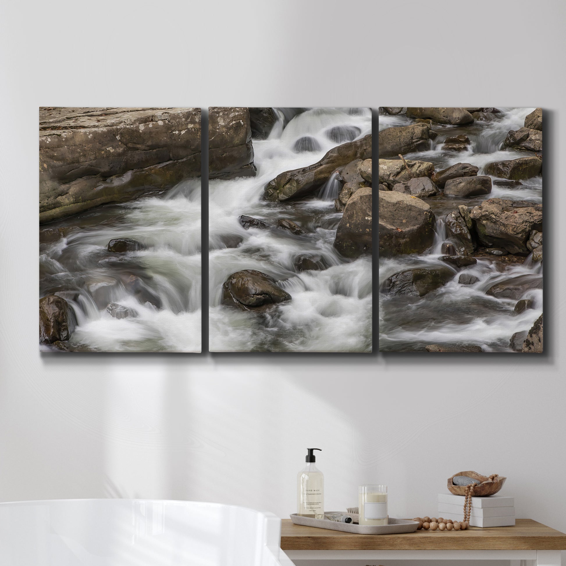 Three-panel canvas wall art featuring rushing river water over rocks, displayed in a bathroom above a bathtub.