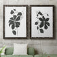 Set of two black barnwood framed canvas prints depicting a coordinated art pair of black leafy vine silhouettes rendered as minimalist botanical prints above a sofa in a modern living room.