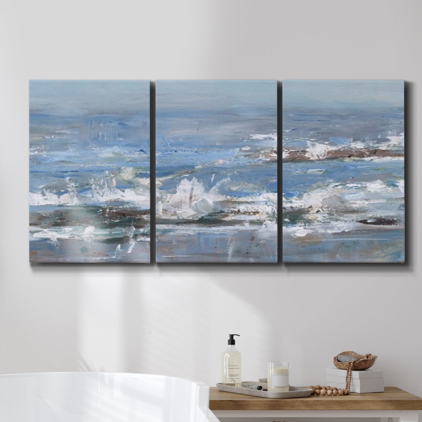 Three-panel canvas wall art featuring overcast coastal waves under grey sky, displayed in a bathroom above a bathtub.