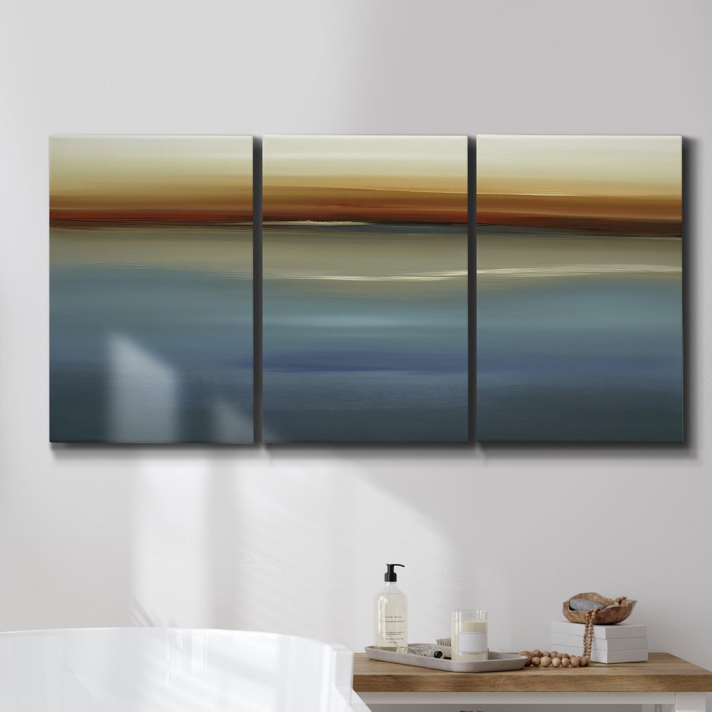 Three-panel canvas wall art featuring horizontal abstract coastal color bands, displayed in a bathroom above a bathtub.