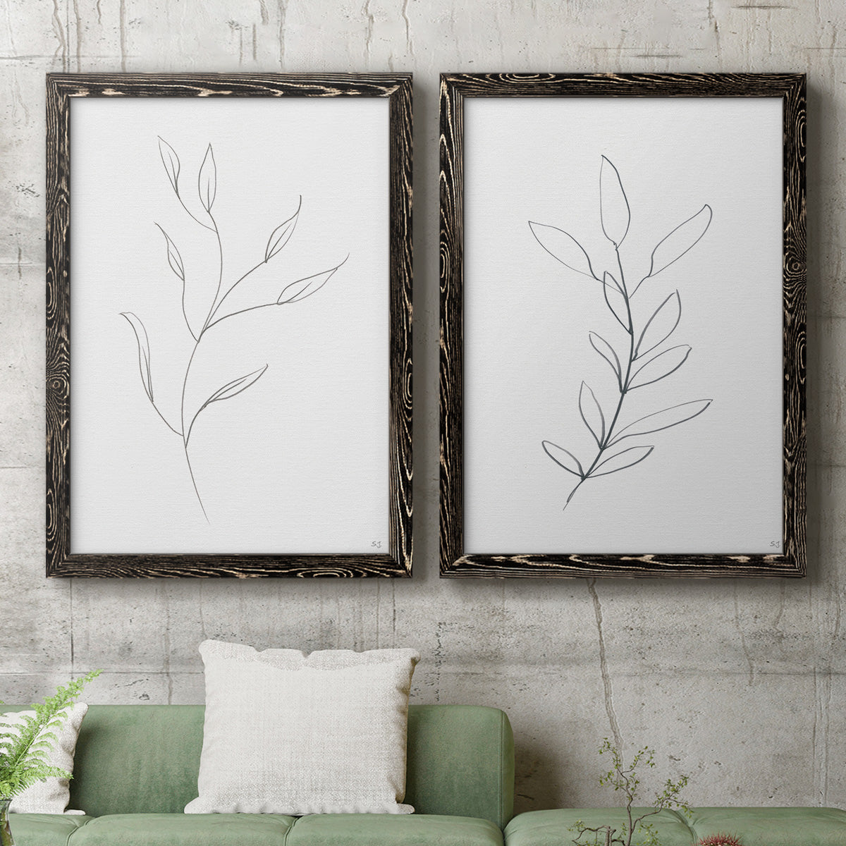 Set of two black barnwood framed canvas prints depicting a coordinated art pair of single continuous-line leafy branches rendered as minimalist botanical line drawings above a sofa in a modern living room.