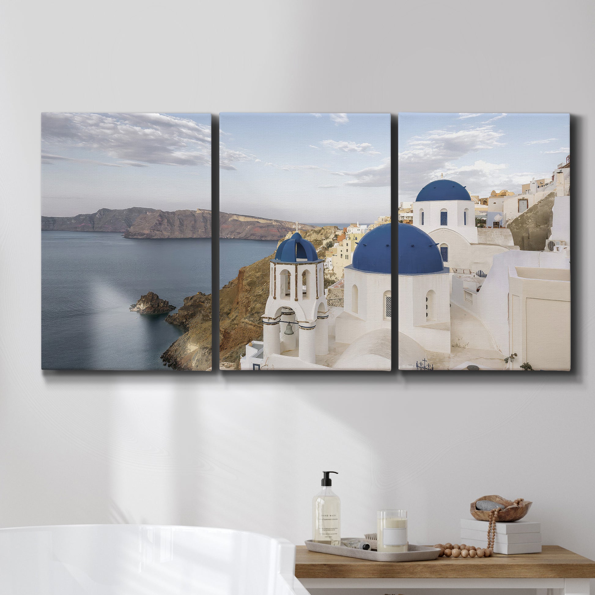 Three-panel canvas wall art featuring white buildings overlooking sea cliffs, displayed in a bathroom above a bathtub.