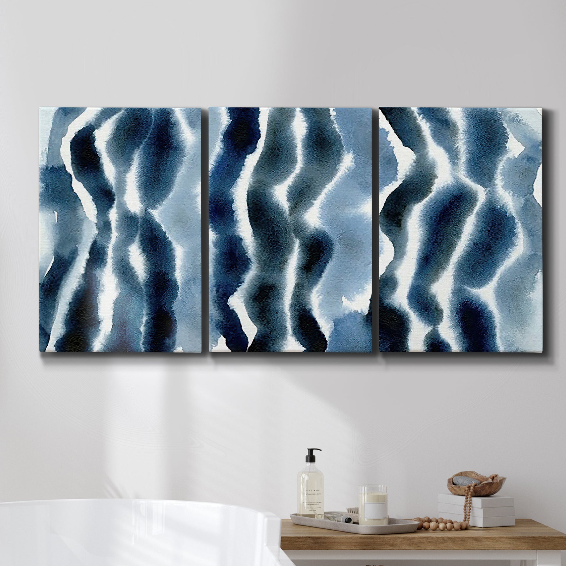 Three-panel canvas wall art featuring flowing blue watercolor wave forms, displayed in a bathroom above a bathtub.