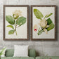Set of two walnut barnwood framed canvas prints depicting a coordinated art pair of white magnolia blossoms and glossy green leaves rendered as traditional botanical paintings above a sofa in a modern living room.