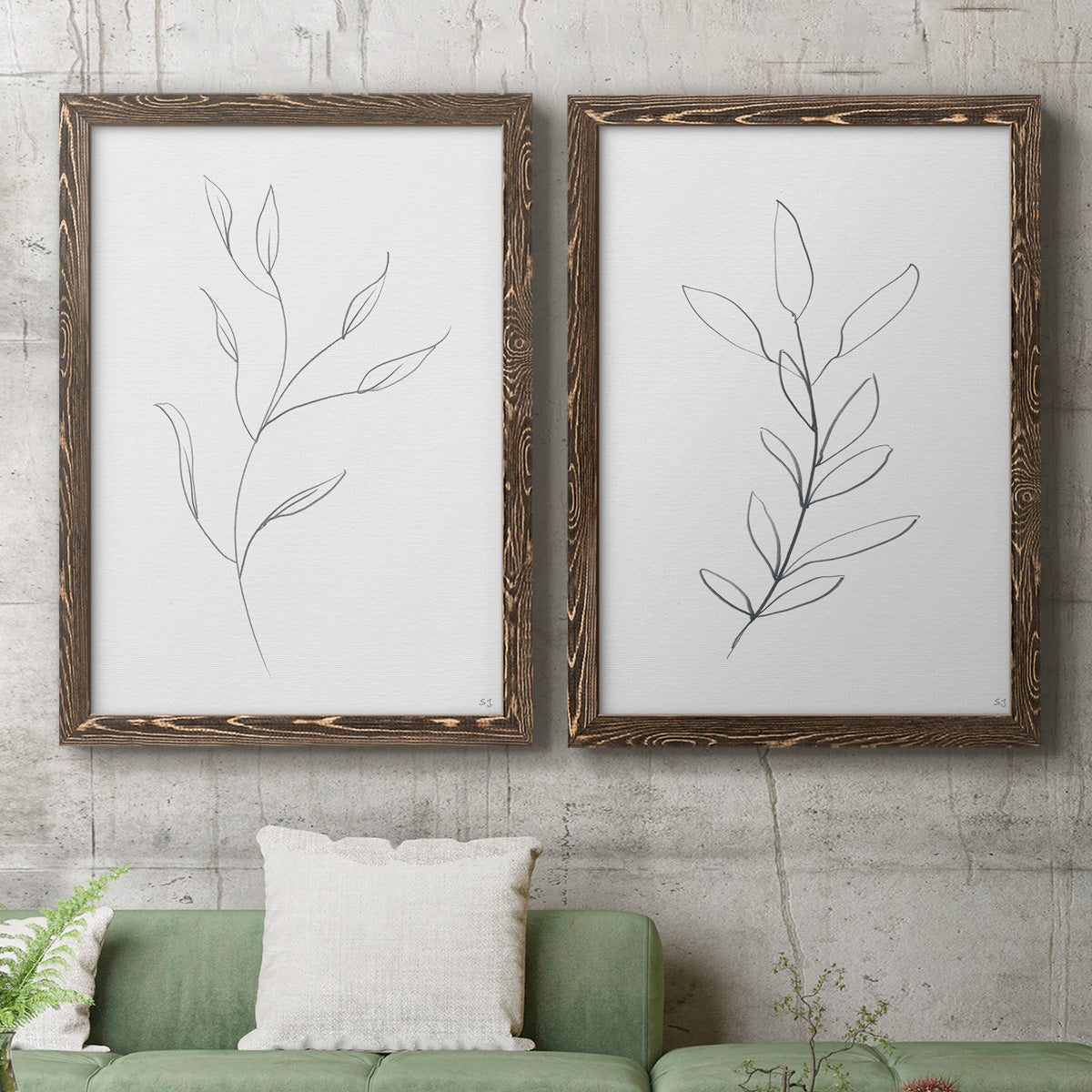 Set of two walnut barnwood framed canvas prints depicting a coordinated art pair of single continuous-line leafy branches rendered as minimalist botanical line drawings above a sofa in a modern living room.