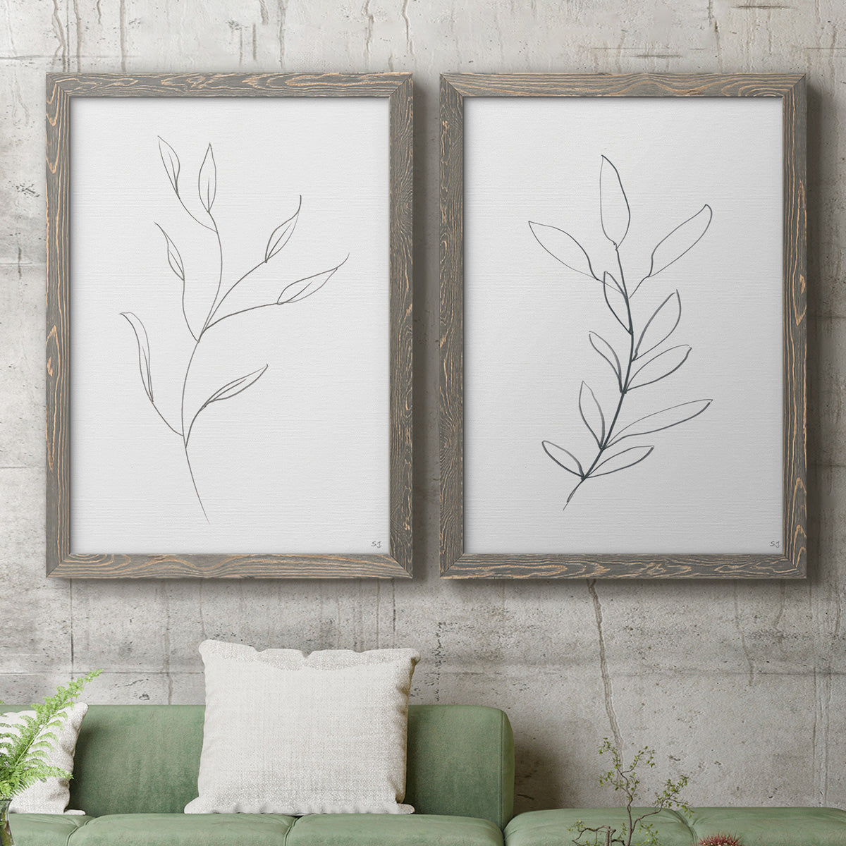 Set of two gray barnwood framed canvas prints depicting a coordinated art pair of single continuous-line leafy branches rendered as minimalist botanical line drawings above a sofa in a modern living room.