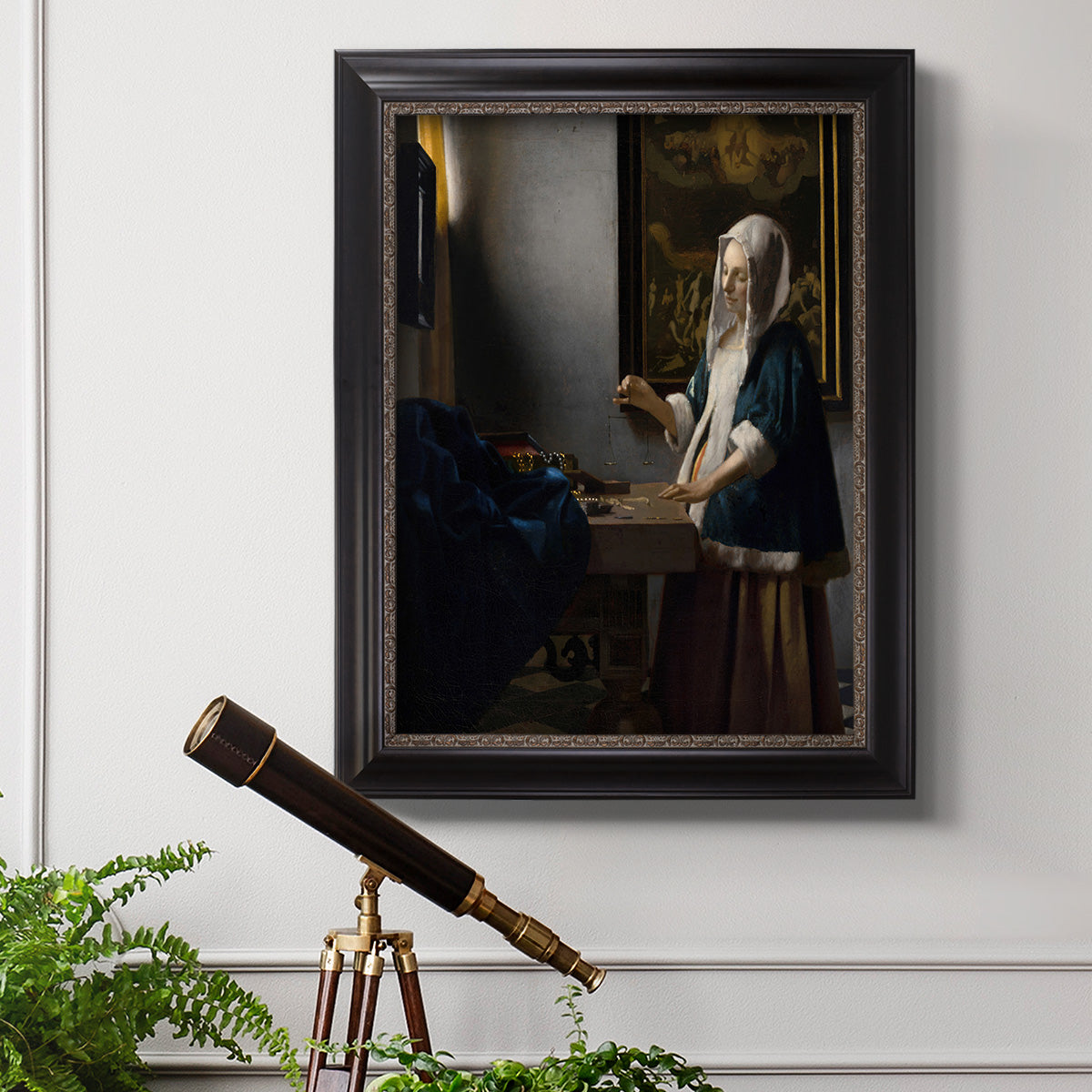 Quiet interior genre scene of a woman with scales by Johannes Vermeer in an ornate black frame with gilded inner detail displayed in a light-toned interior setting.