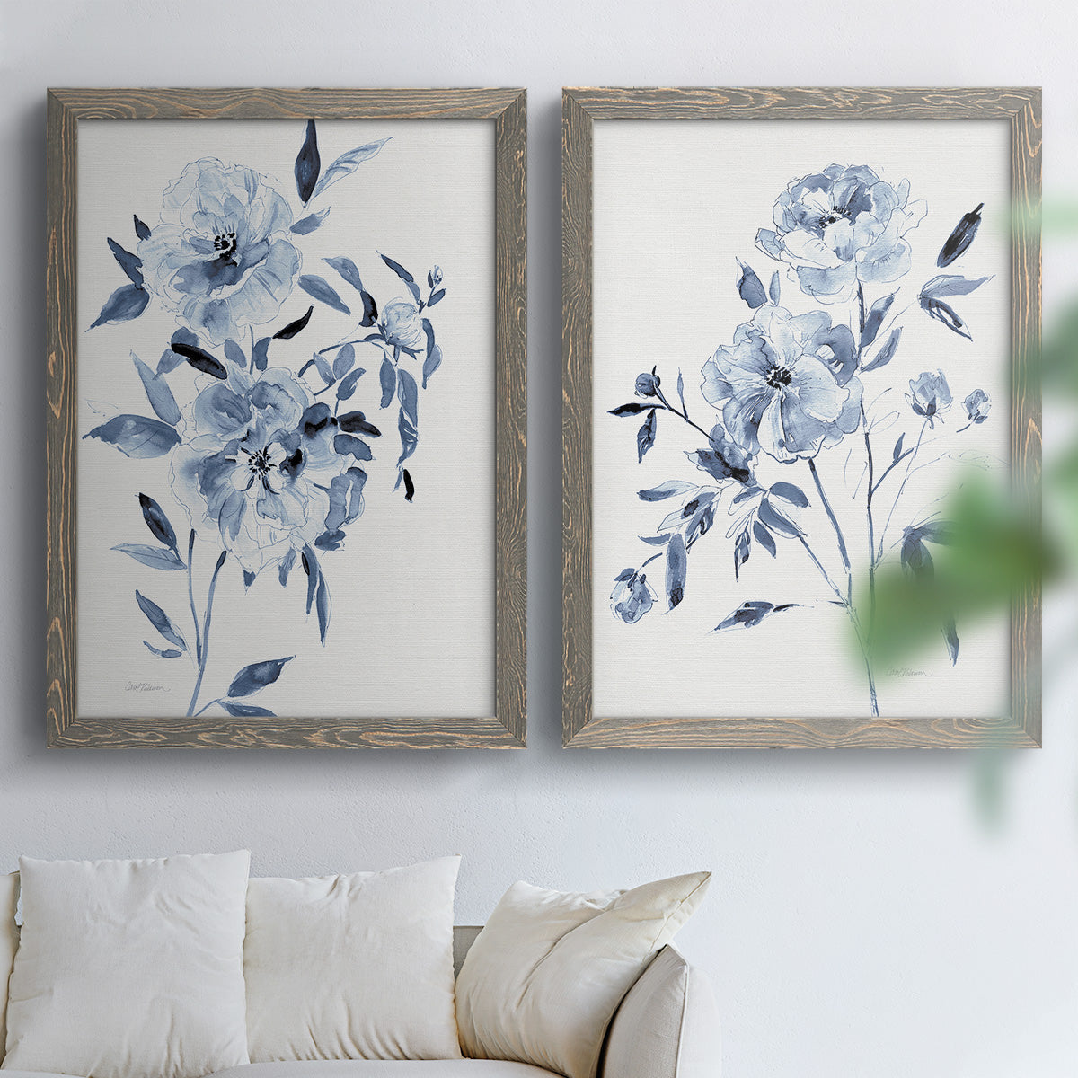 Set of two gray barnwood framed canvas prints depicting indigo blossoms, gestural blue leaves, and airy white space arranged across two complementary compositions above a sofa in a bright living space.