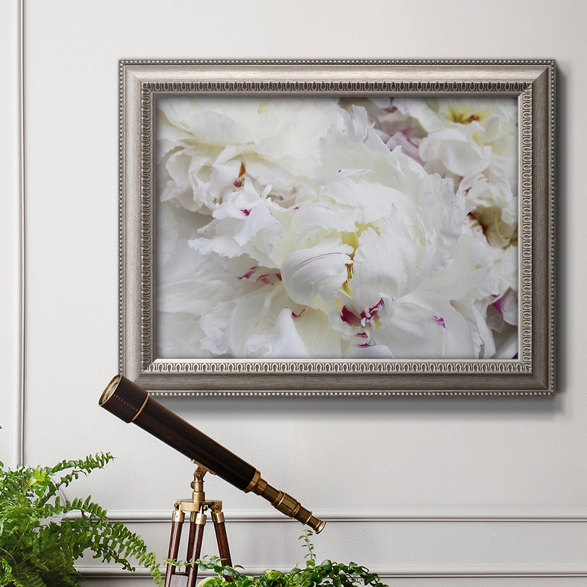 White peonies in bloom in an ornate vintage silver frame displayed in a light-toned interior setting.