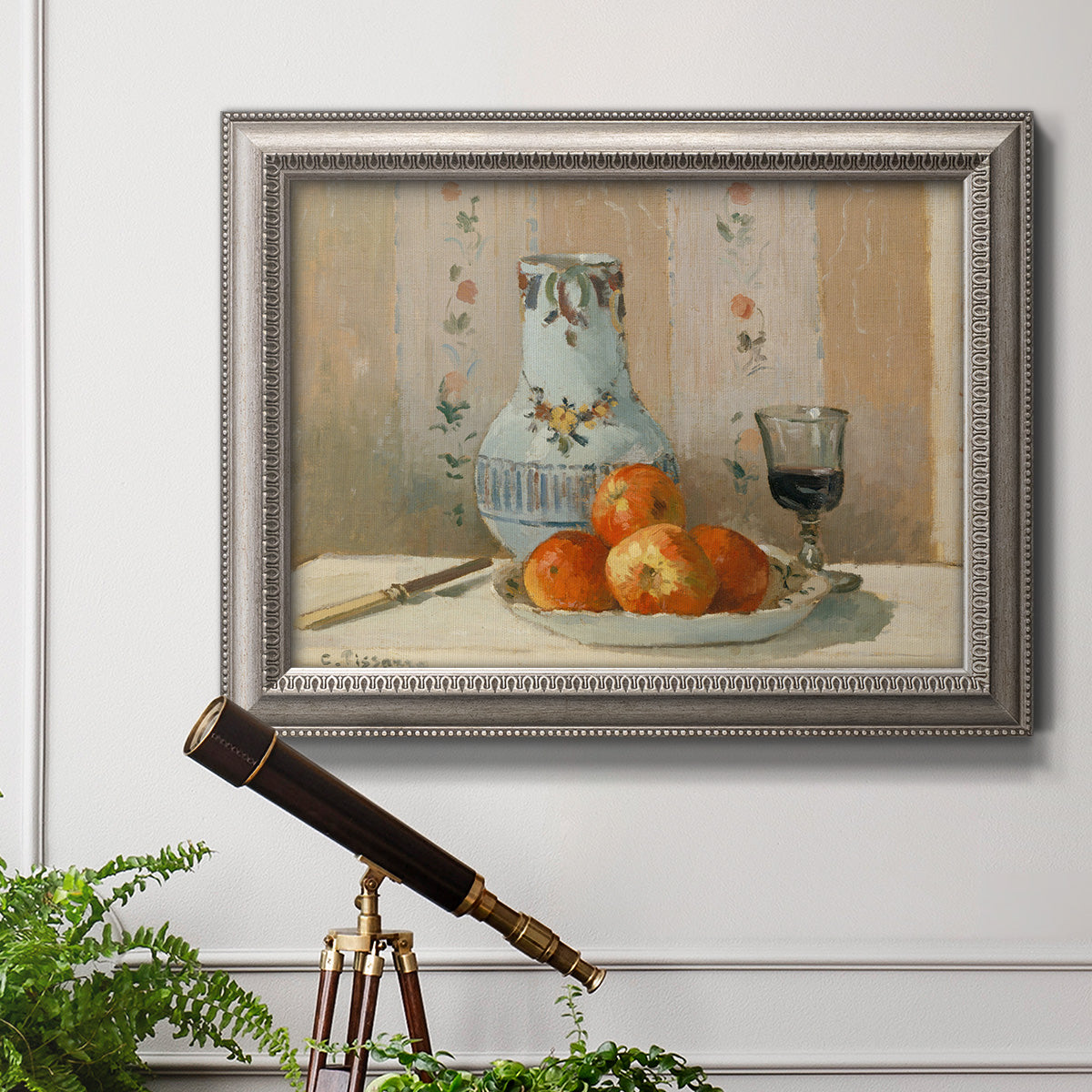 Impressionist still life painting with apples and a ceramic pitcher in an ornate vintage silver frame displayed in a light-toned interior setting.