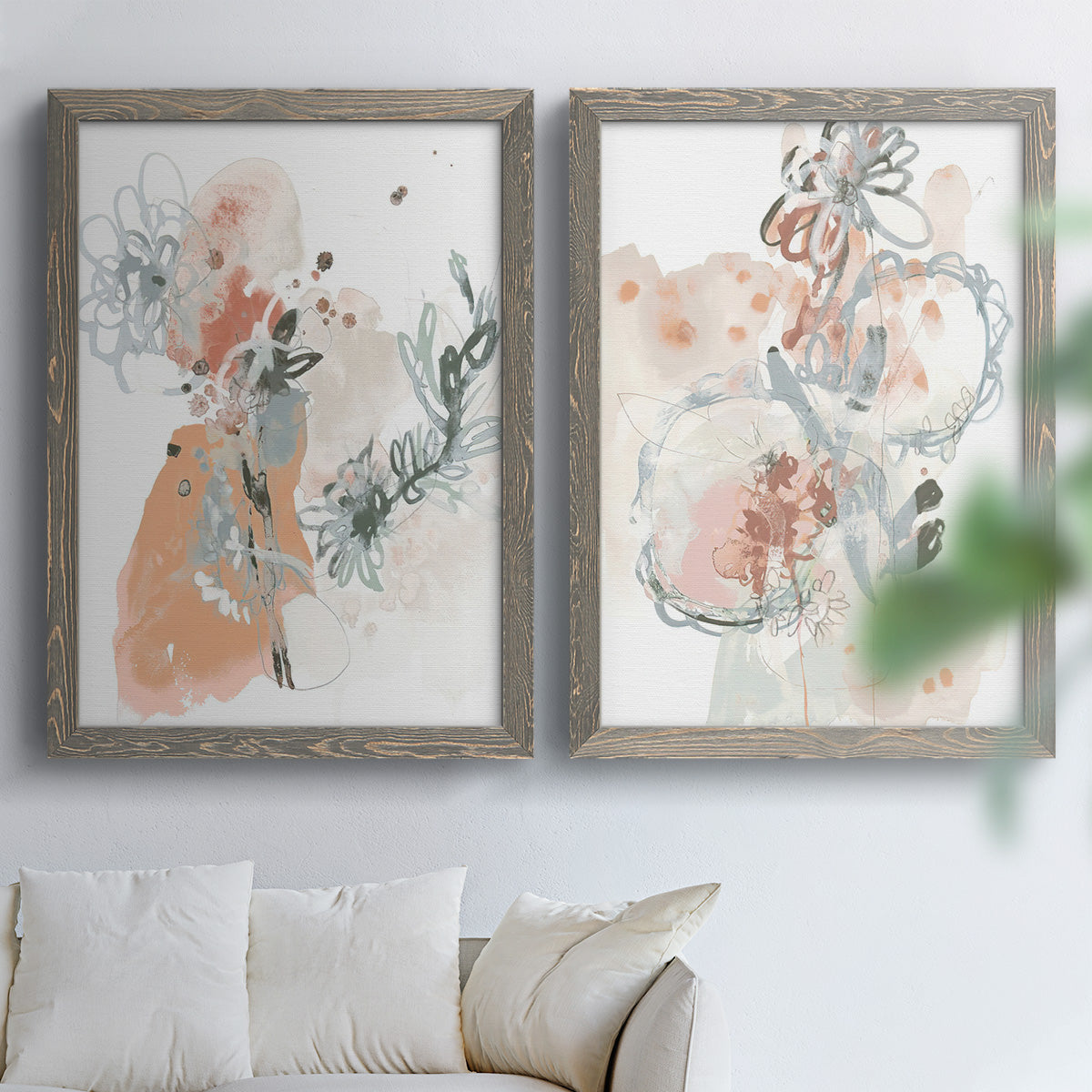 Set of two gray barnwood framed canvas prints depicting scribbled petals, translucent washes, and drifting stems arranged across two complementary compositions above a sofa in a bright living space.