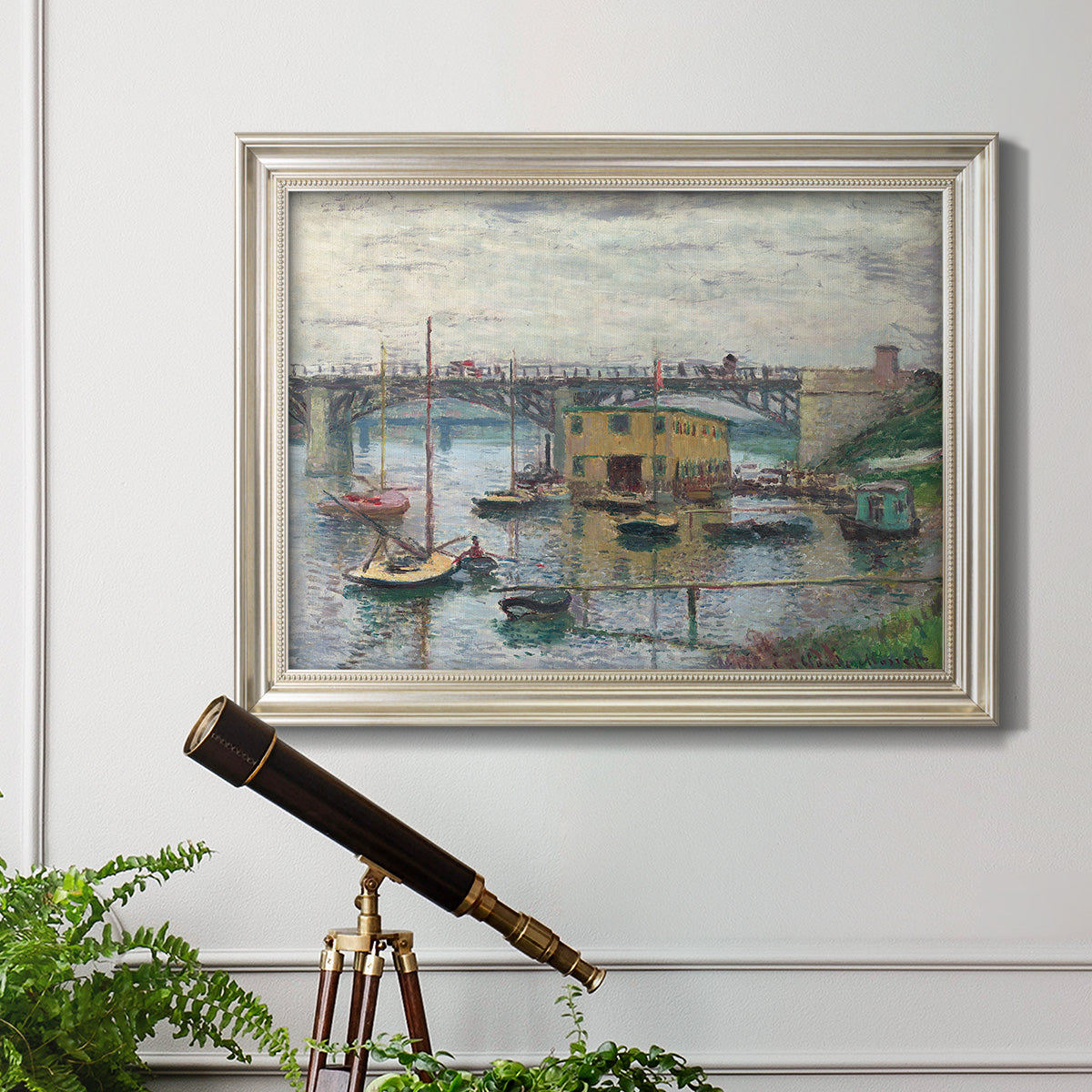 Impressionist river bridge scene on an overcast day by Claude Monet in an ornate champagne frame displayed in a light-toned interior setting.
