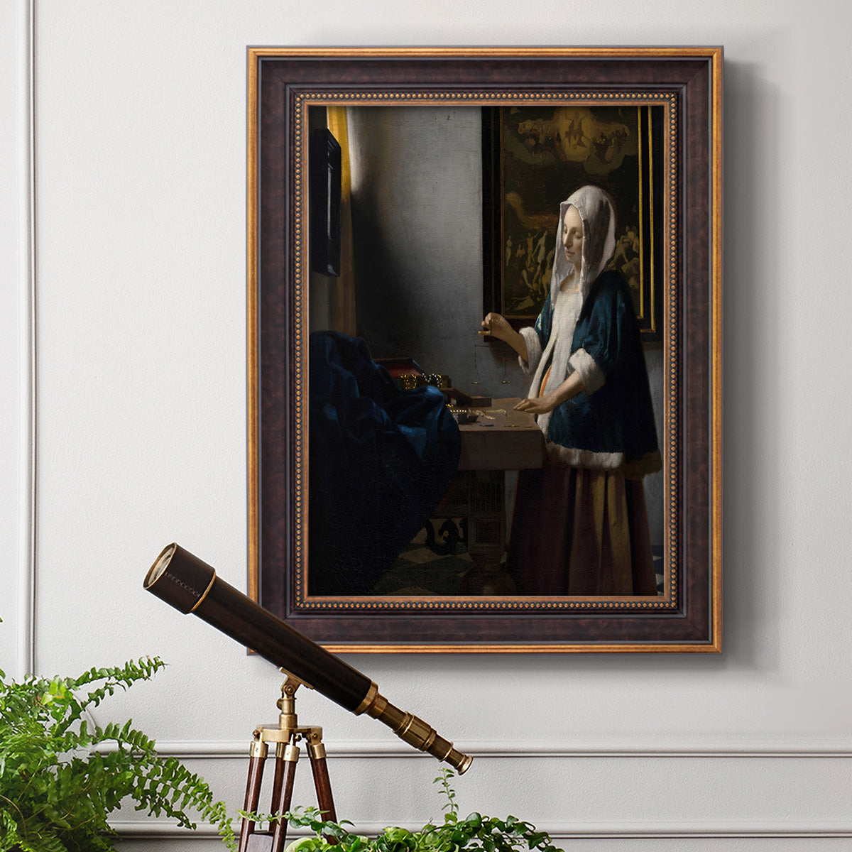 Quiet interior genre scene of a woman with scales by Johannes Vermeer in an ornate bronzed mahogany wood frame displayed in a light-toned interior setting.
