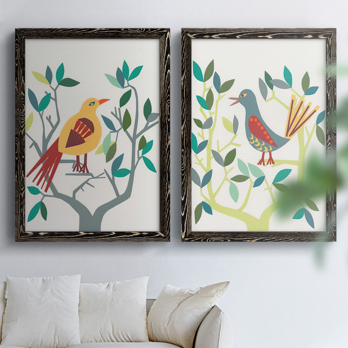 Set of two black barnwood framed canvas prints depicting red yellow blue plumage, simplified leaves, and playful flat shapes arranged across two complementary compositions above a sofa in a bright living space.