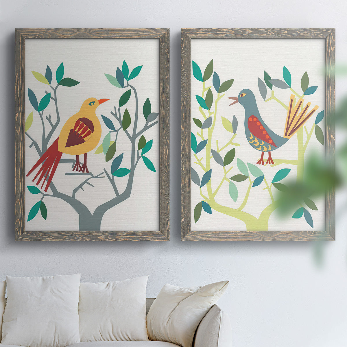 Set of two gray barnwood framed canvas prints depicting red yellow blue plumage, simplified leaves, and playful flat shapes arranged across two complementary compositions above a sofa in a bright living space.