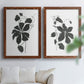 Set of two maple barnwood framed canvas prints depicting bold dark leaves, curving stems, and clean white grounds arranged across two complementary compositions above a sofa in a bright living space.