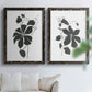 Set of two black barnwood framed canvas prints depicting bold dark leaves, curving stems, and clean white grounds arranged across two complementary compositions above a sofa in a bright living space.