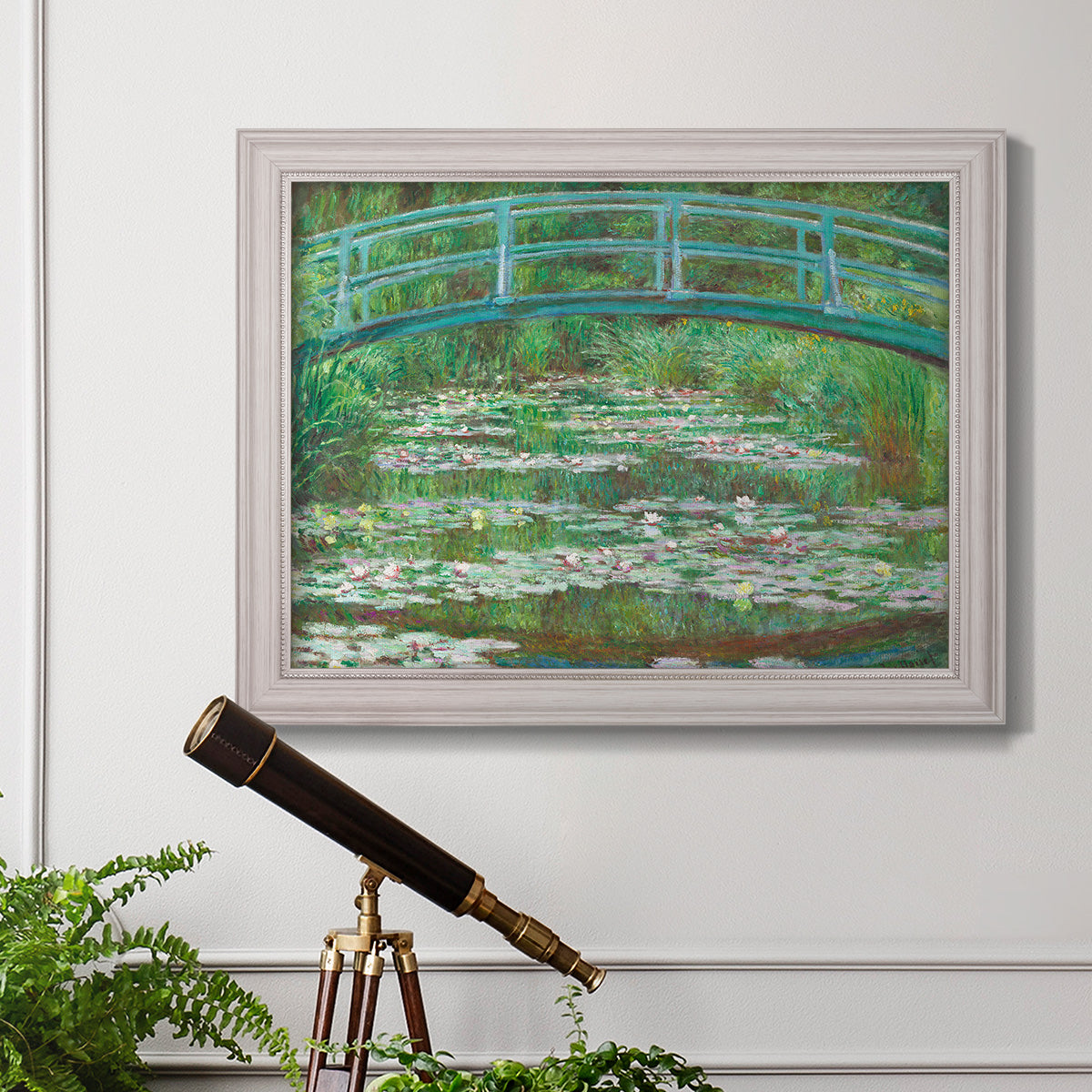 Impressionist garden scene with Japanese bridge and water lilies by Claude Monet in an ornate weathered white frame displayed in a light-toned interior setting.