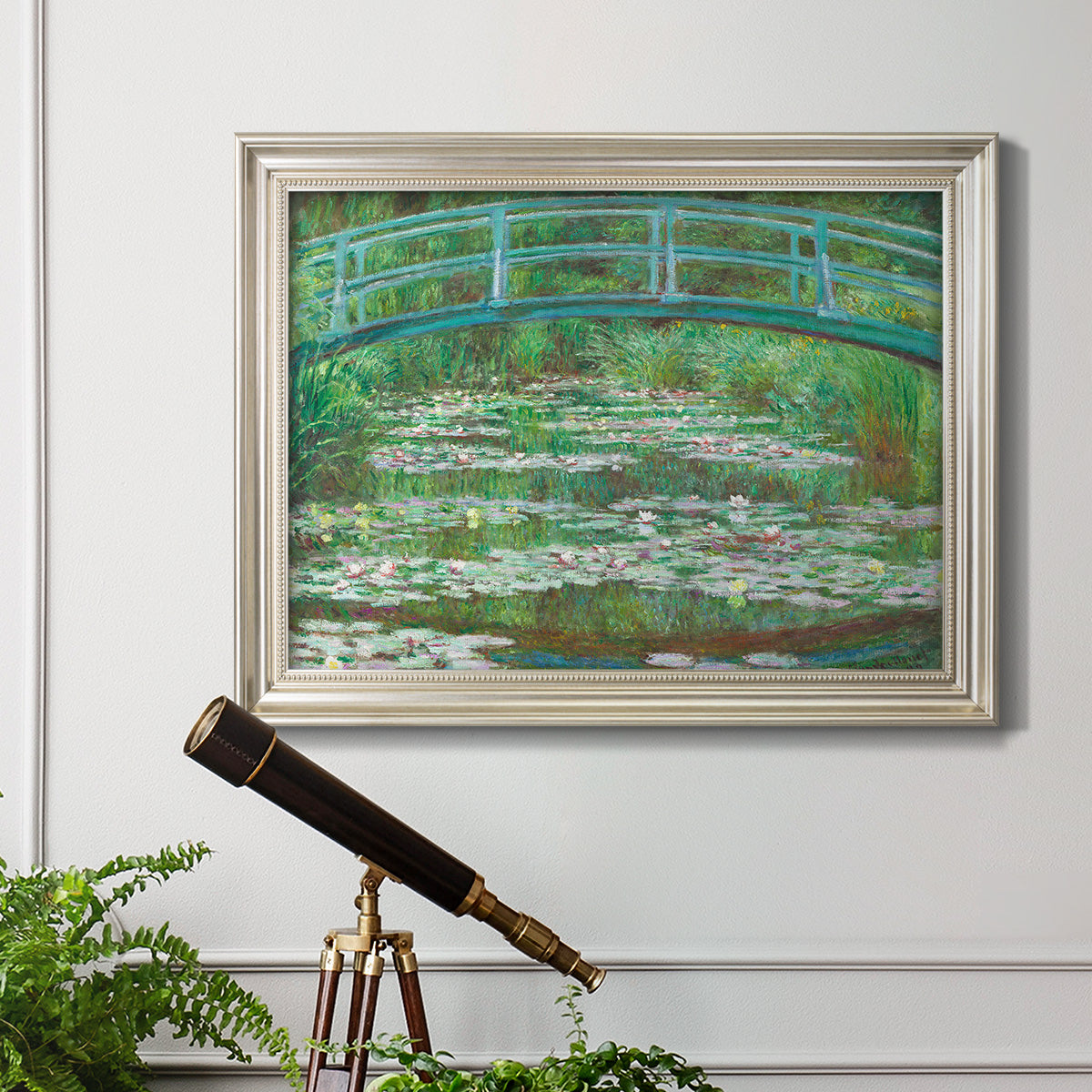 Impressionist garden scene with Japanese bridge and water lilies by Claude Monet in an ornate champagne frame displayed in a light-toned interior setting.