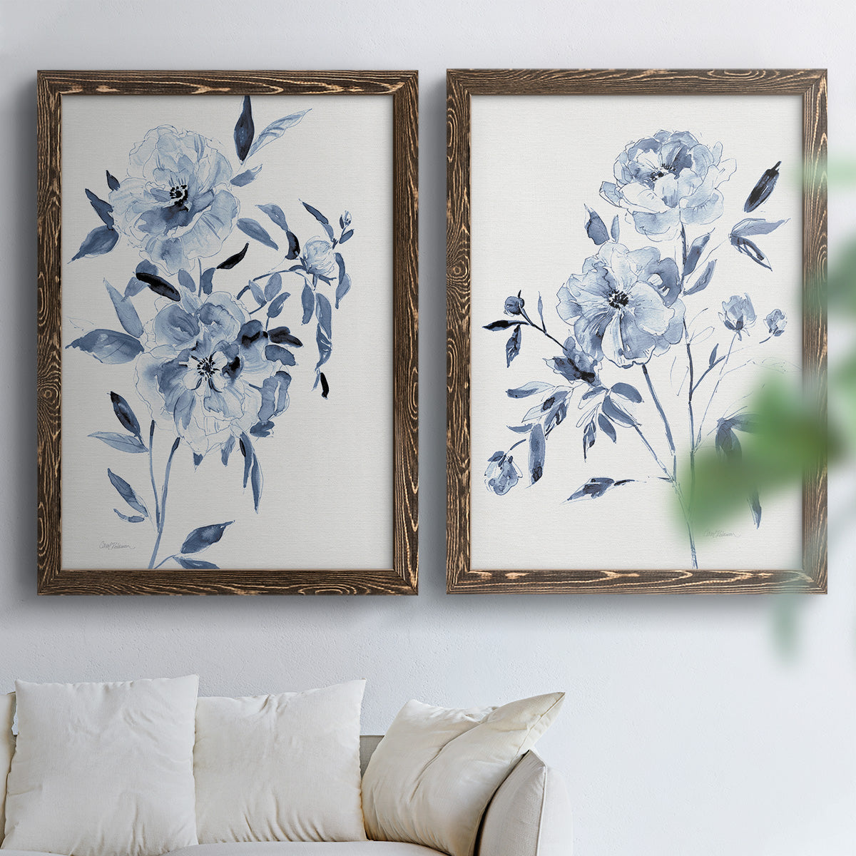 Set of two walnut barnwood framed canvas prints depicting indigo blossoms, gestural blue leaves, and airy white space arranged across two complementary compositions above a sofa in a bright living space.