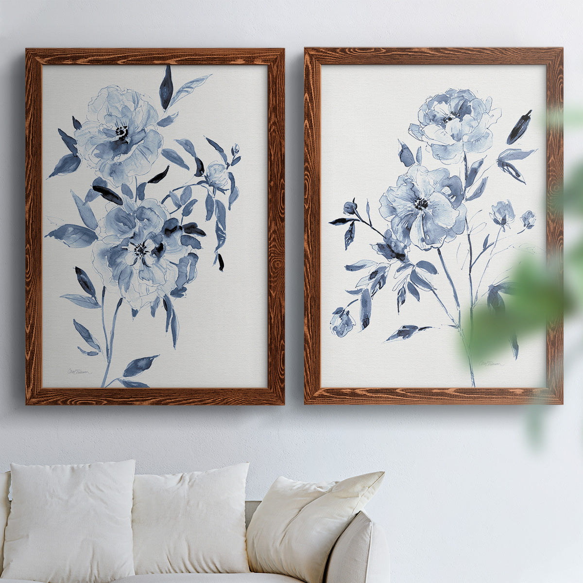 Set of two maple barnwood framed canvas prints depicting indigo blossoms, gestural blue leaves, and airy white space arranged across two complementary compositions above a sofa in a bright living space.