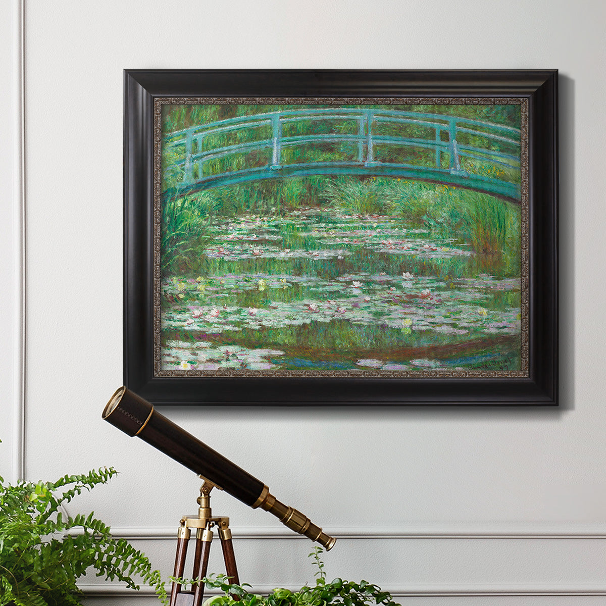 Impressionist garden scene with Japanese bridge and water lilies by Claude Monet in an ornate black frame with gilded inner detail displayed in a light-toned interior setting.