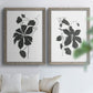 Set of two gray barnwood framed canvas prints depicting bold dark leaves, curving stems, and clean white grounds arranged across two complementary compositions above a sofa in a bright living space.