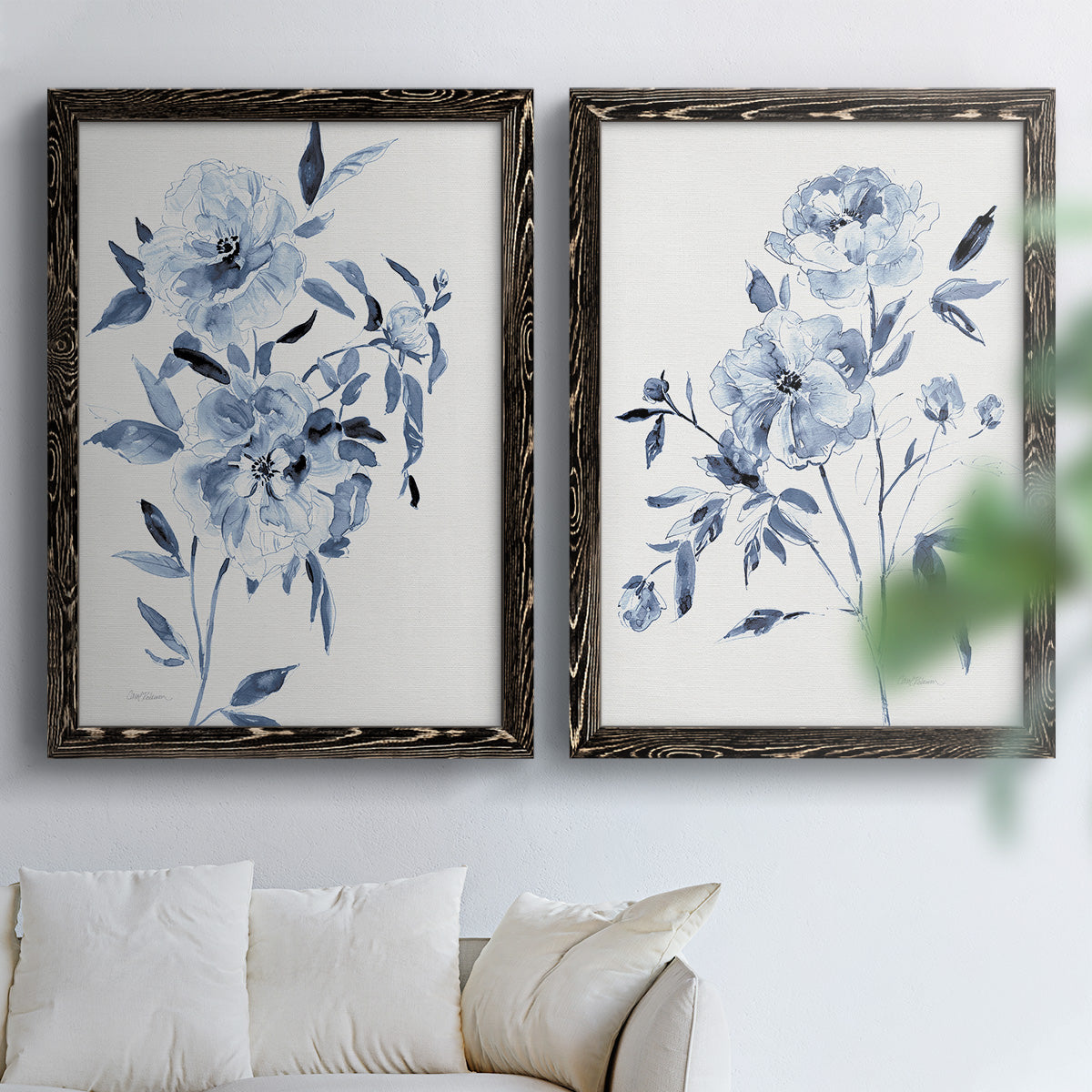 Set of two black barnwood framed canvas prints depicting indigo blossoms, gestural blue leaves, and airy white space arranged across two complementary compositions above a sofa in a bright living space.