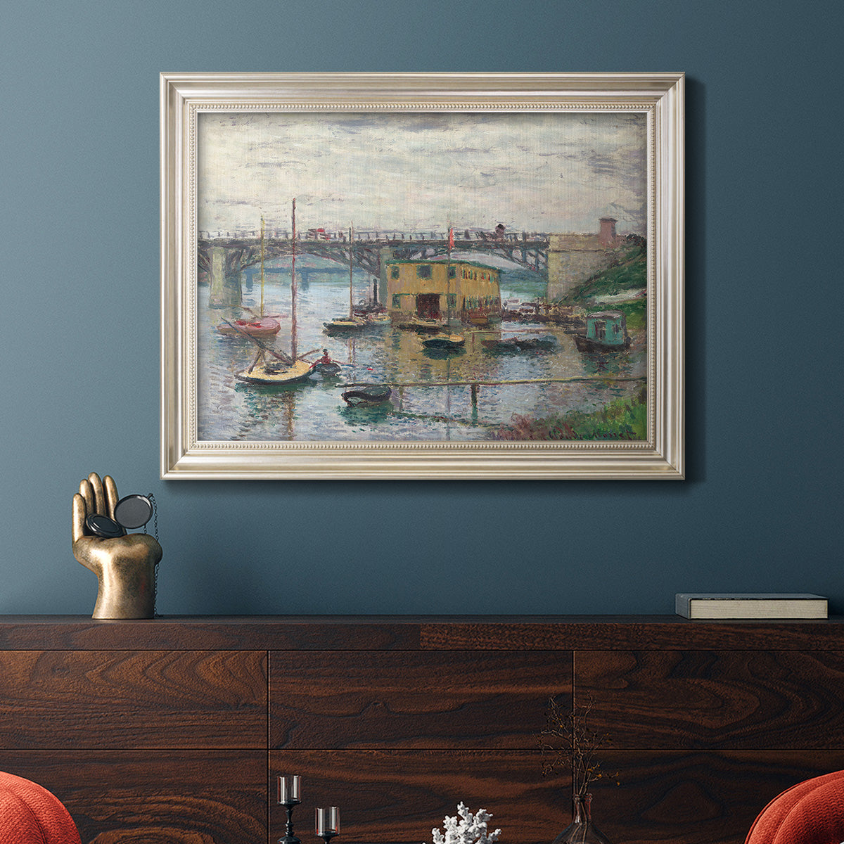 Impressionist river bridge scene on an overcast day by Claude Monet in an ornate champagne frame displayed in a dark-toned interior setting.