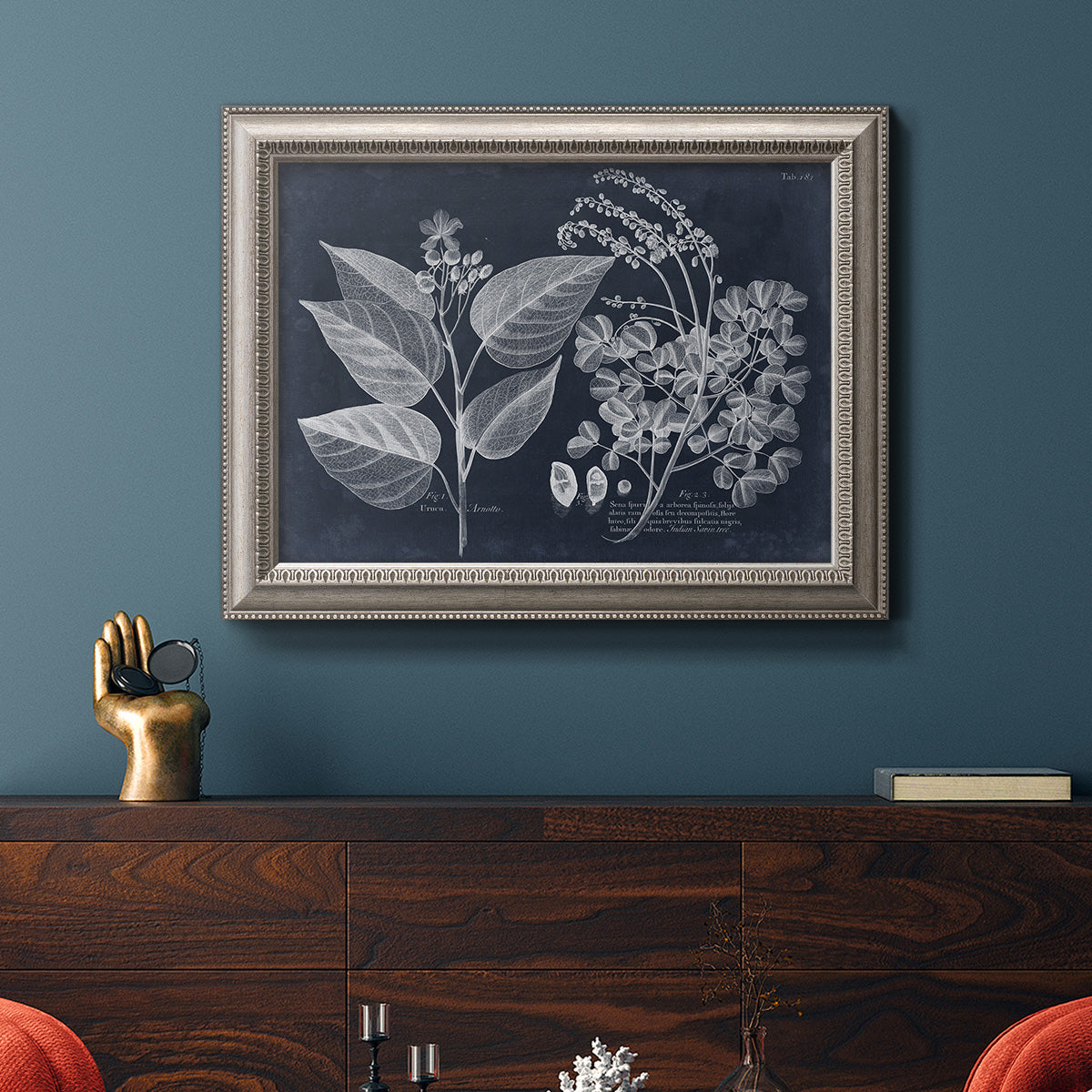 Decorative botanical foliage drawing on navy background in an ornate vintage silver frame displayed in a dark-toned interior setting.