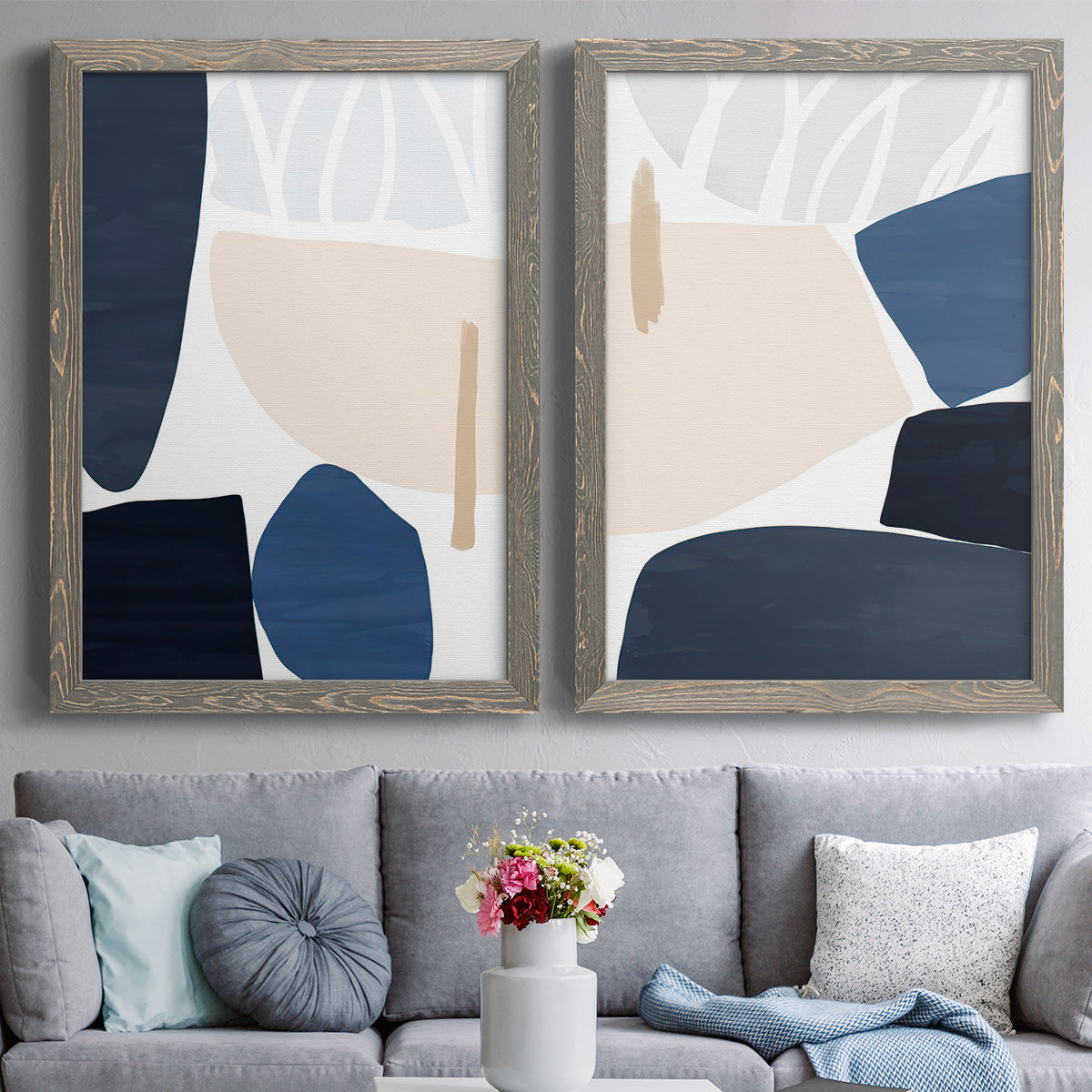 Set of two gray barnwood framed canvas prints depicting paired minimalist color-block abstracts with rounded dark forms, pale sand fields, and clean modern edges above a sofa in a contemporary living room.