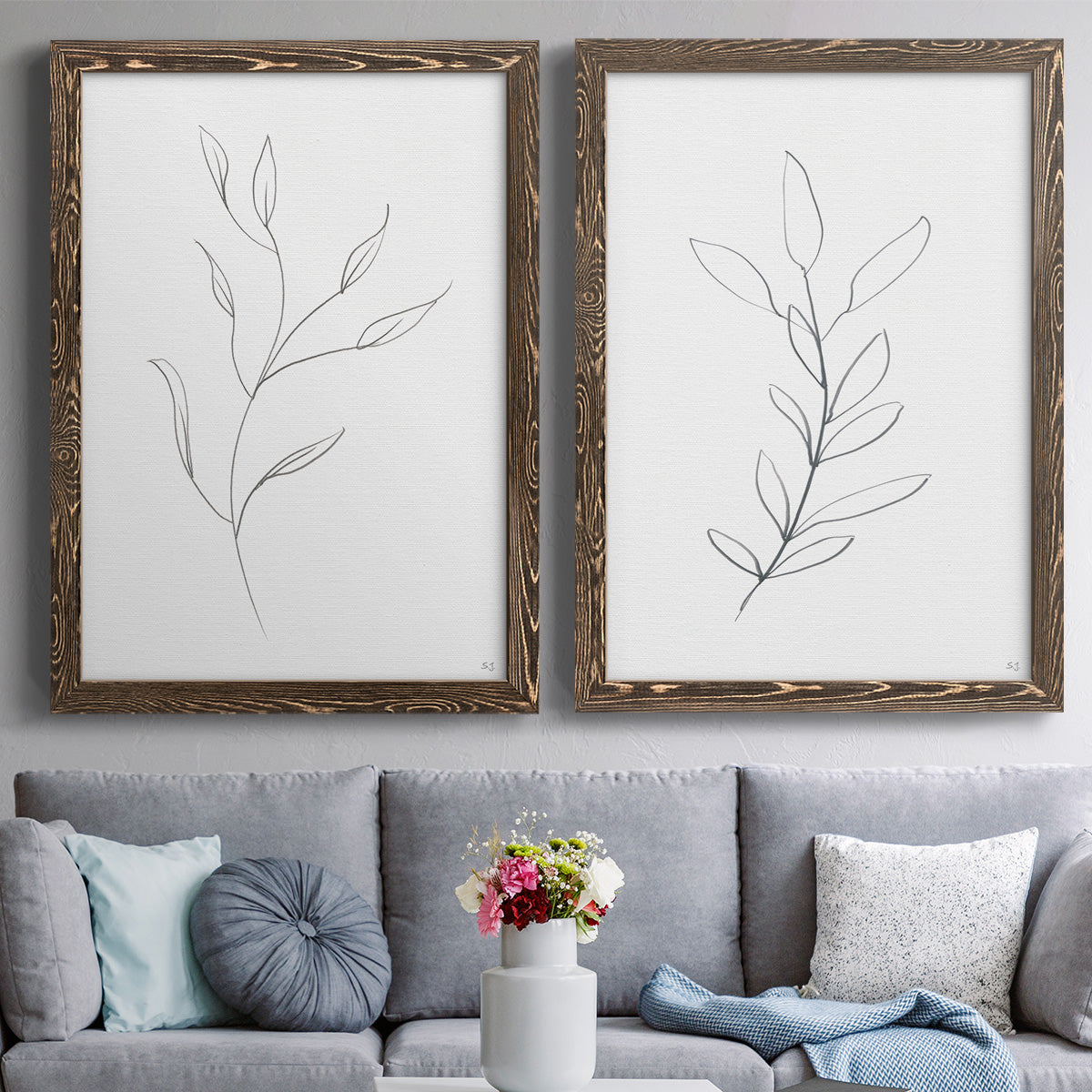 Set of two walnut barnwood framed canvas prints depicting paired minimalist botanical line drawings with thin gray contours, sparse stems, and clean white fields above a sofa in a contemporary living room.