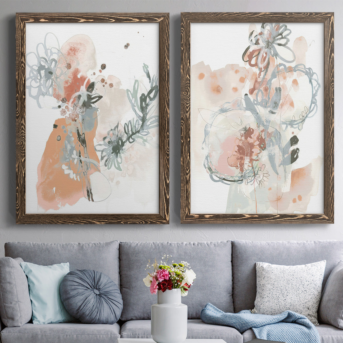 Set of two walnut barnwood framed canvas prints depicting paired abstract watercolor botanicals with scribbled petals, translucent washes, and drifting stems above a sofa in a contemporary living room.