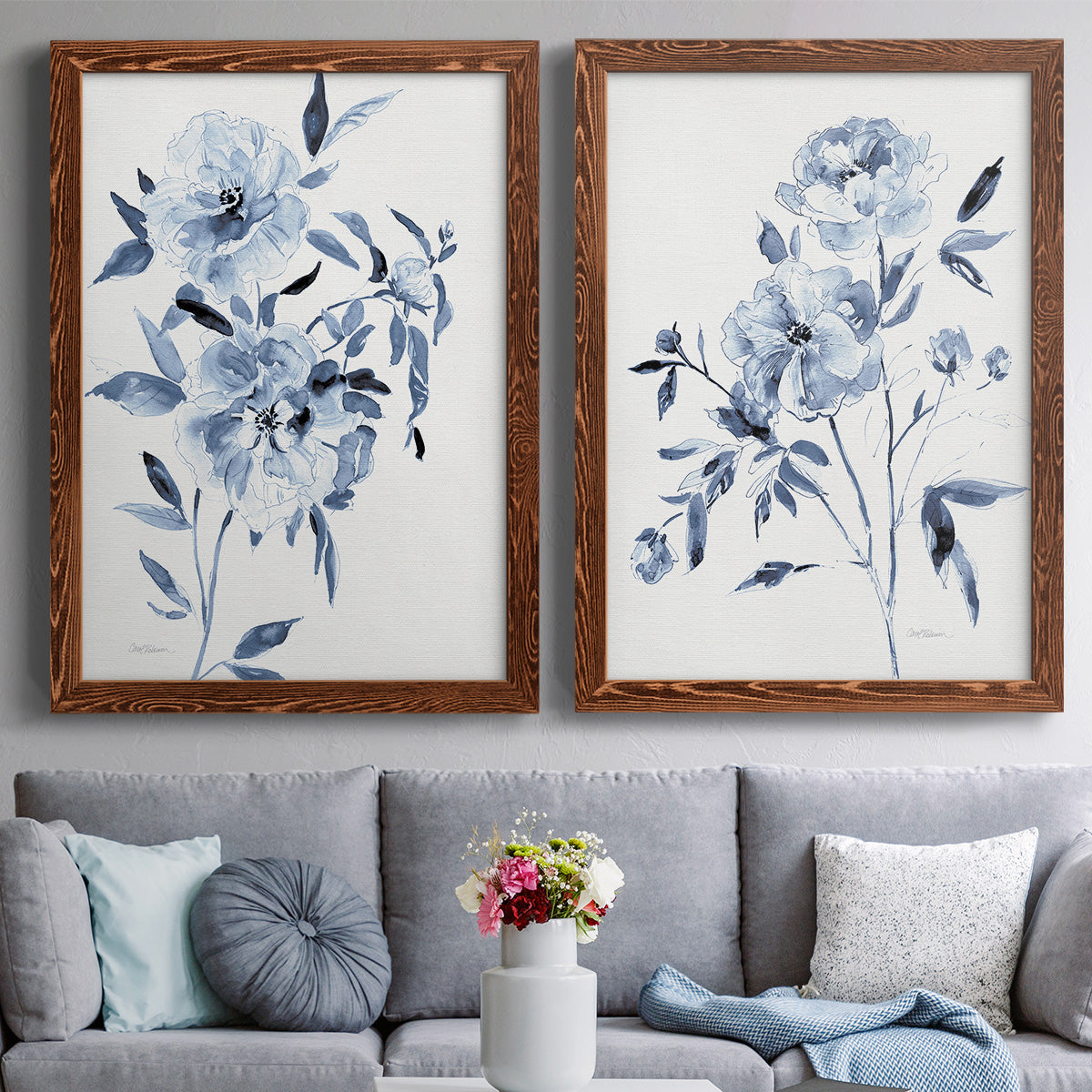 Set of two maple barnwood framed canvas prints depicting paired expressive watercolor botanical paintings with indigo blossoms, gestural blue leaves, and airy white space above a sofa in a contemporary living room.