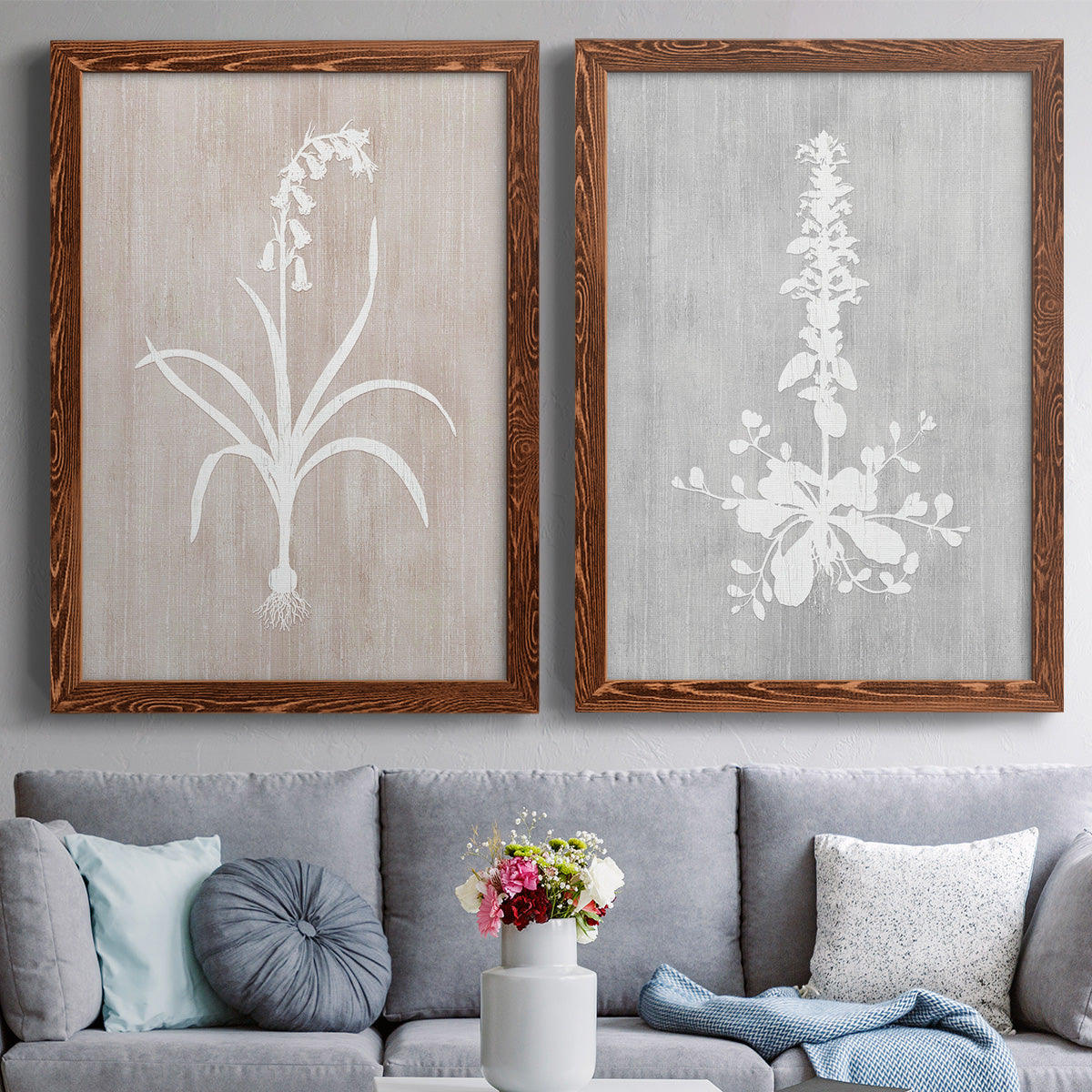 Set of two maple barnwood framed canvas prints depicting paired rustic minimalist botanical prints with delicate stems, leaves, and blossoms against gray taupe and pale blue panels above a sofa in a contemporary living room.
