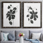 Set of two black barnwood framed canvas prints depicting paired minimalist botanical prints with bold dark leaves, curving stems, and clean white grounds above a sofa in a contemporary living room.